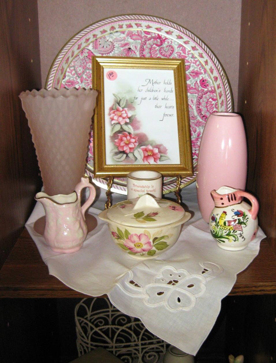 Shelf display of pink ceramics and a framed floral print on a lace-covered table.