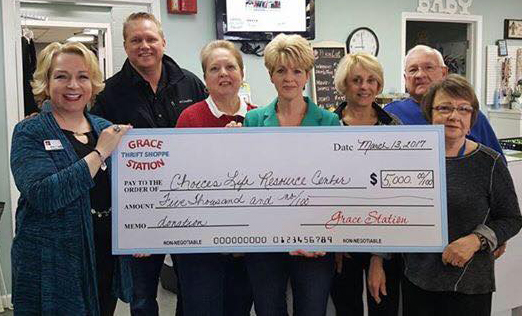 Eight people stand in a shop holding a large ceremonial check, smiling for the camera. The oversized check indicates a $5,000 donation to a charity, suggesting a group donation event.